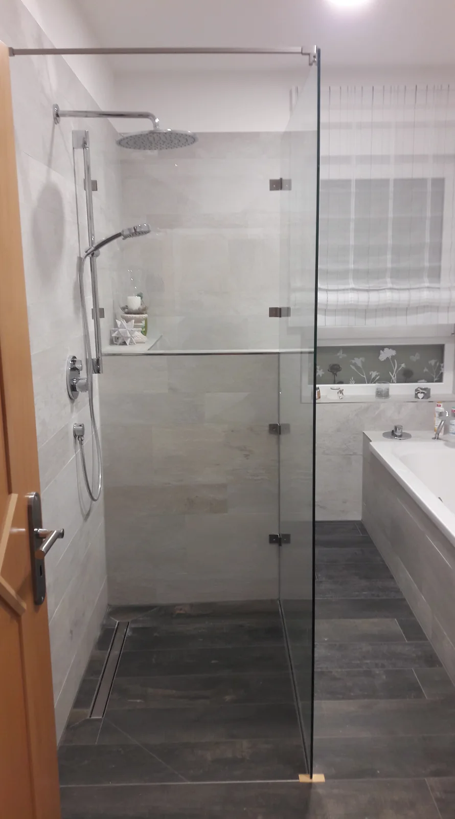 Duschabtrennung aus Glas Gläserne Duschabtrennung mit Metallbeschlägen und oberer Stabilisierungsstange vor einer gefliesten Dusche.