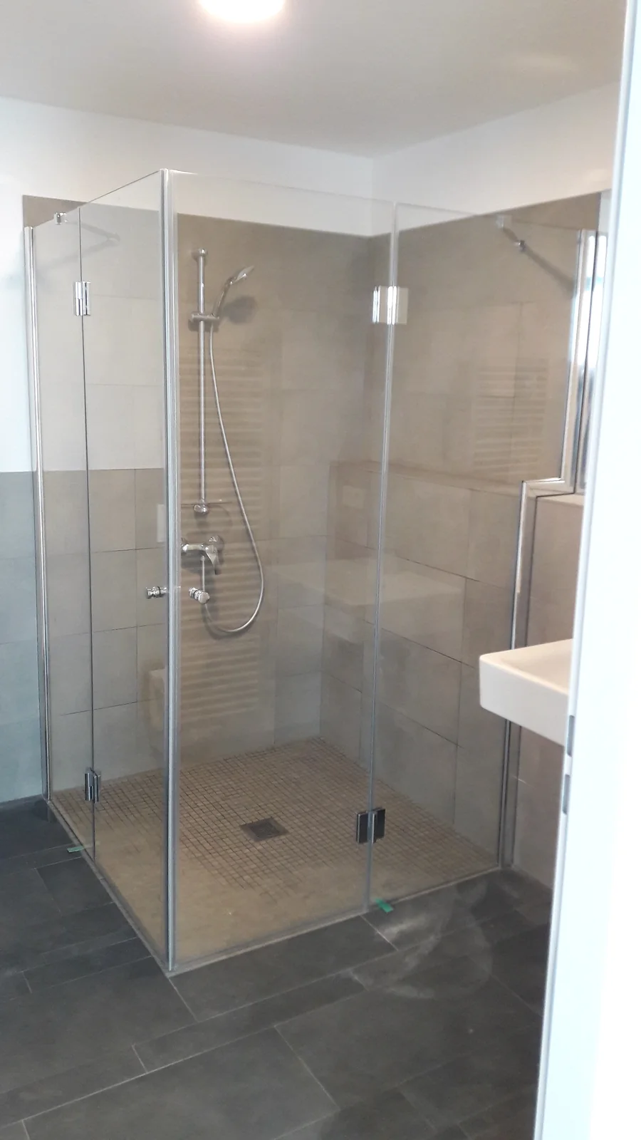 Glasdusche mit angepasstem Seitenteil Glasdusche mit Metallbeschlägen, Mosaikboden und abgestuftem Seitenteil neben dem Waschtisch.
