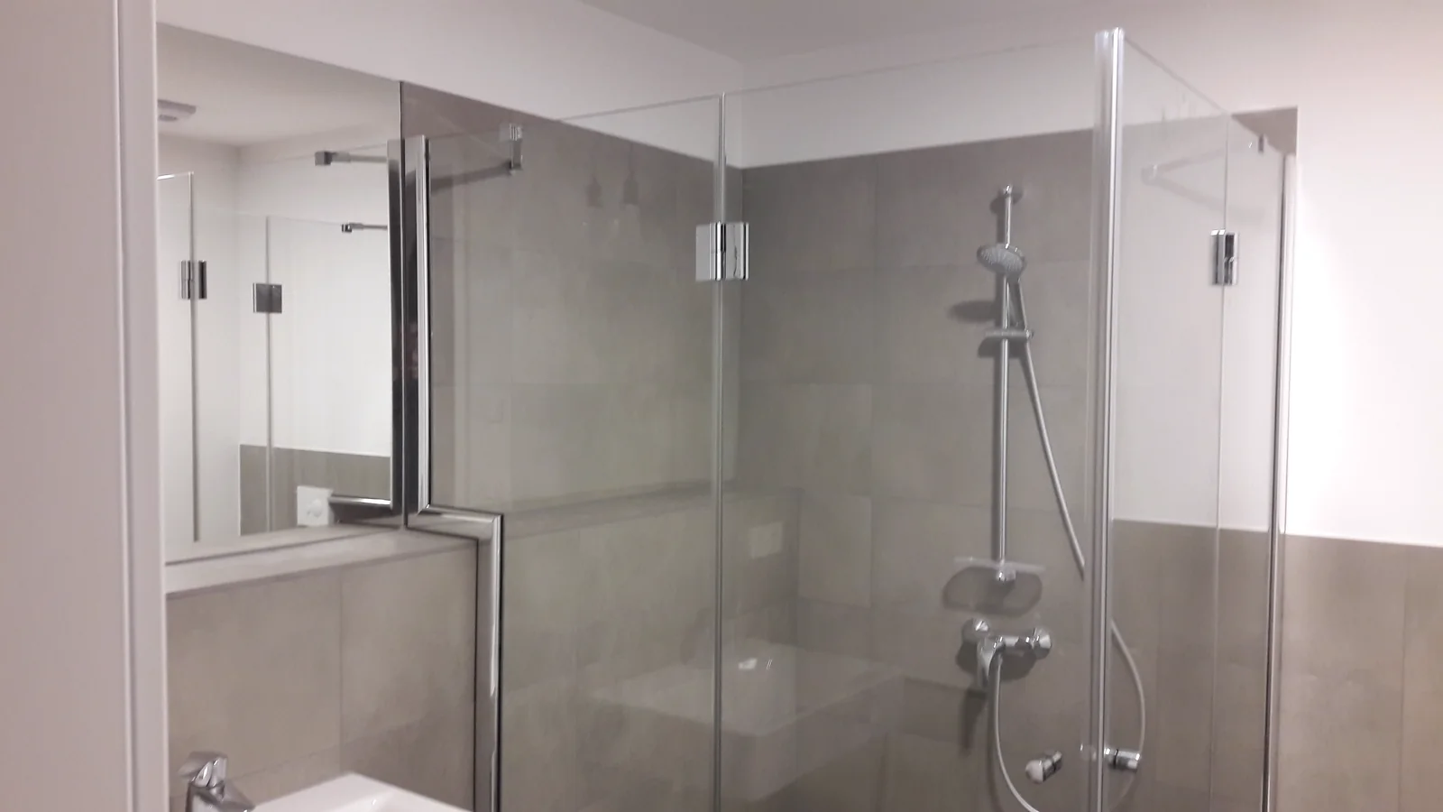 Glasdusche neben dem Waschtisch Gläserne Duschabtrennung mit Metallbeschlägen neben einem Waschtisch und Wandspiegel.